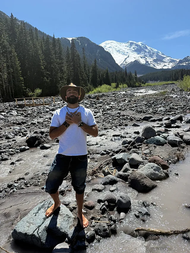 Standing in a river with hands over his heart, Mount Rainier behind