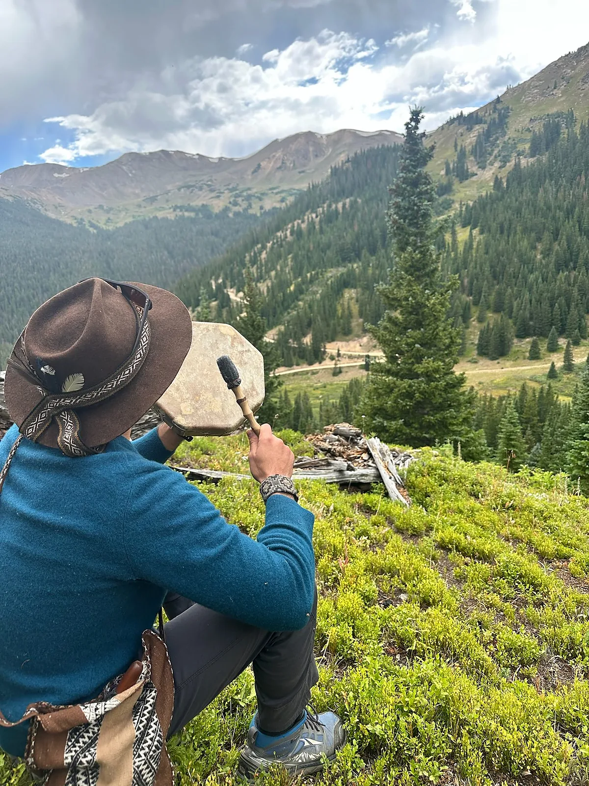 Izzy seated on a mountainside, seen from behind, drum in his hands, looking out